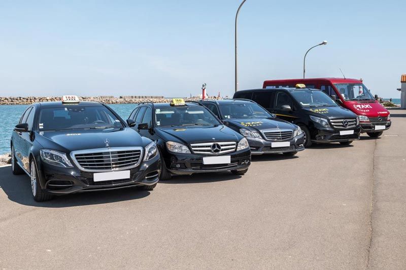 Plusieurs taxis noirs sont stationnés le long du front de mer. Une camionnette rouge ferme la file.