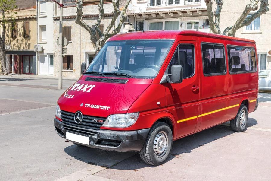Une camionnette Mercedes-Benz Sprinter rouge portant l'inscription « TAXI » est stationnée dans la rue, devant des immeubles.