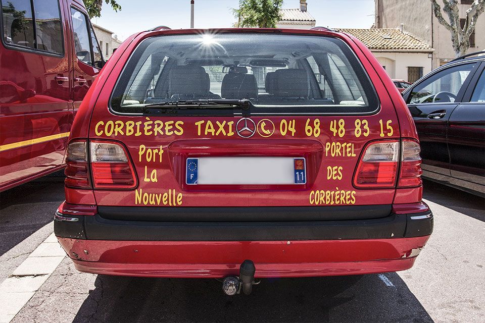 Taxi rouge avec l'inscription « Corbières Taxi » et un numéro de téléphone à l'arrière, stationné dans une rue.