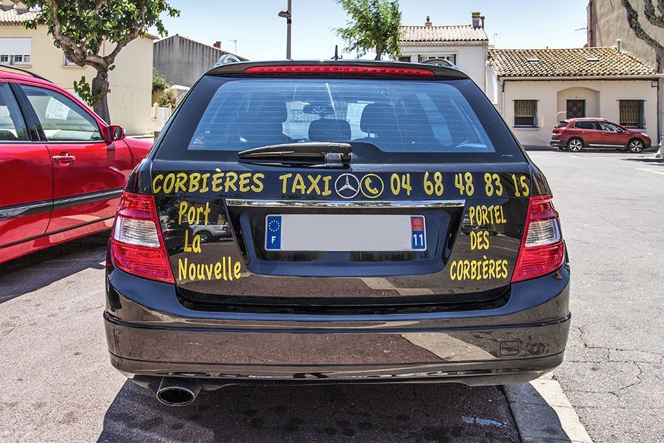 Taxi noir avec l'inscription « Corbières Taxi » et le numéro de téléphone, stationné devant les immeubles.