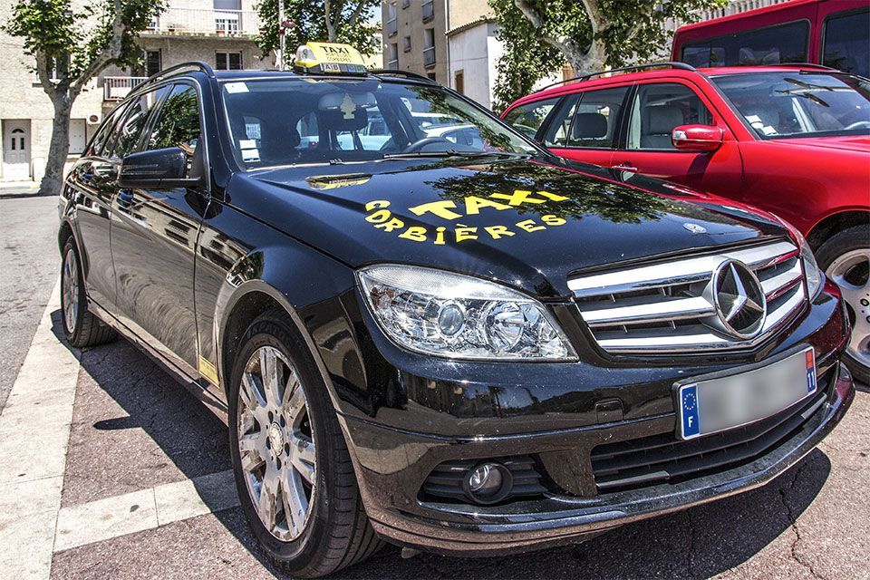 Un taxi Mercedes noir avec un panneau « taxi » est stationné dans une rue à côté d'une voiture rouge.