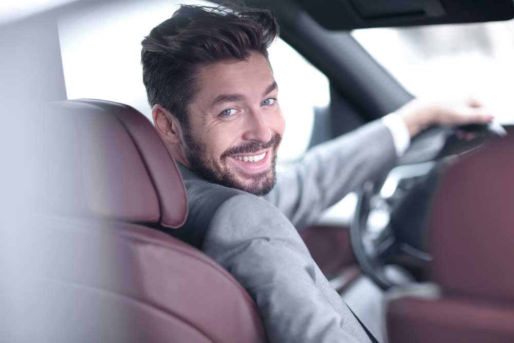 Un homme en costume gris sourit au volant d'une voiture ; l'intérieur en cuir rouge est visible.