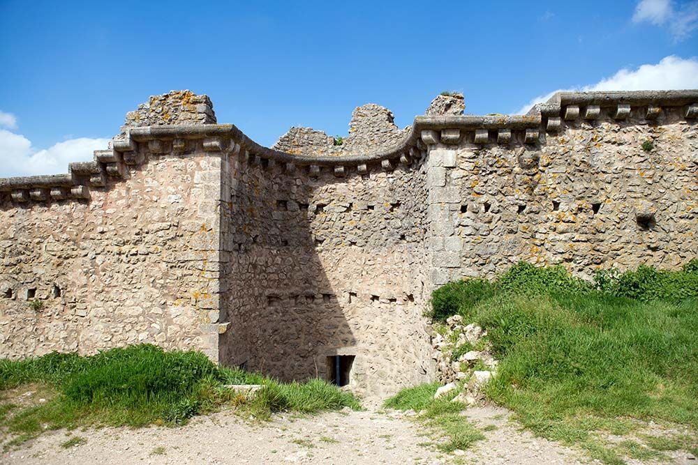 Murs de château en pierre avec créneaux, petites fenêtres et porte sous un ciel bleu.