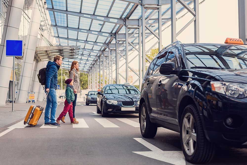 Une famille traverse un passage piéton devant un terminal d'aéroport ; un taxi est visible au premier plan.