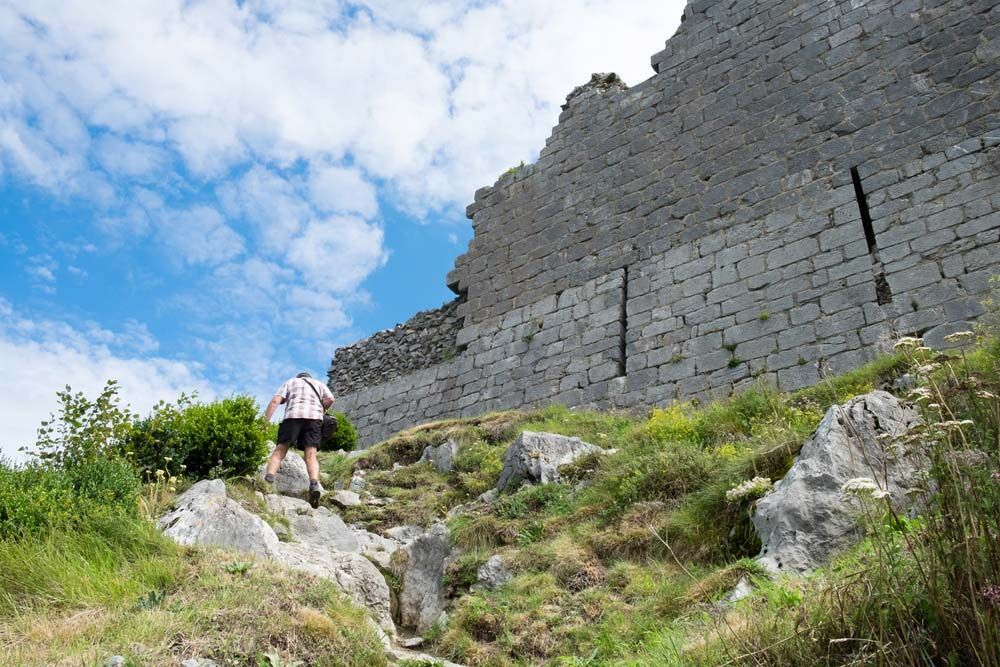 Une personne gravissant un sentier rocailleux vers un grand mur de pierre menant à un château, sous un ciel bleu partiellement nuageux.