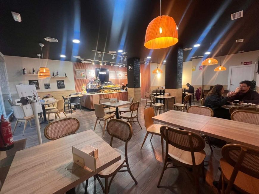 Interior de una cafetería con mesas, sillas y luces naranjas colgantes. Dos personas están sentadas en una mesa.