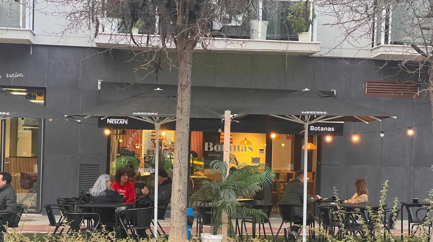 Exterior del restaurante con mesas al aire libre bajo sombrillas. La gente cena en mesas, con un edificio al fondo.