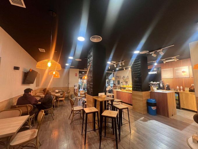 Interior de una cafetería con mesas, sillas y varias personas sentadas. Techo negro, madera y tonos beige.