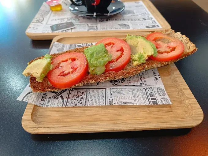 Rodajas de aguacate y tomate sobre pan, servidas en una bandeja de madera con una taza de café de fondo.