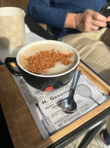 Taza de café con canela en una bandeja, al lado de una persona en un entorno de cafetería.
