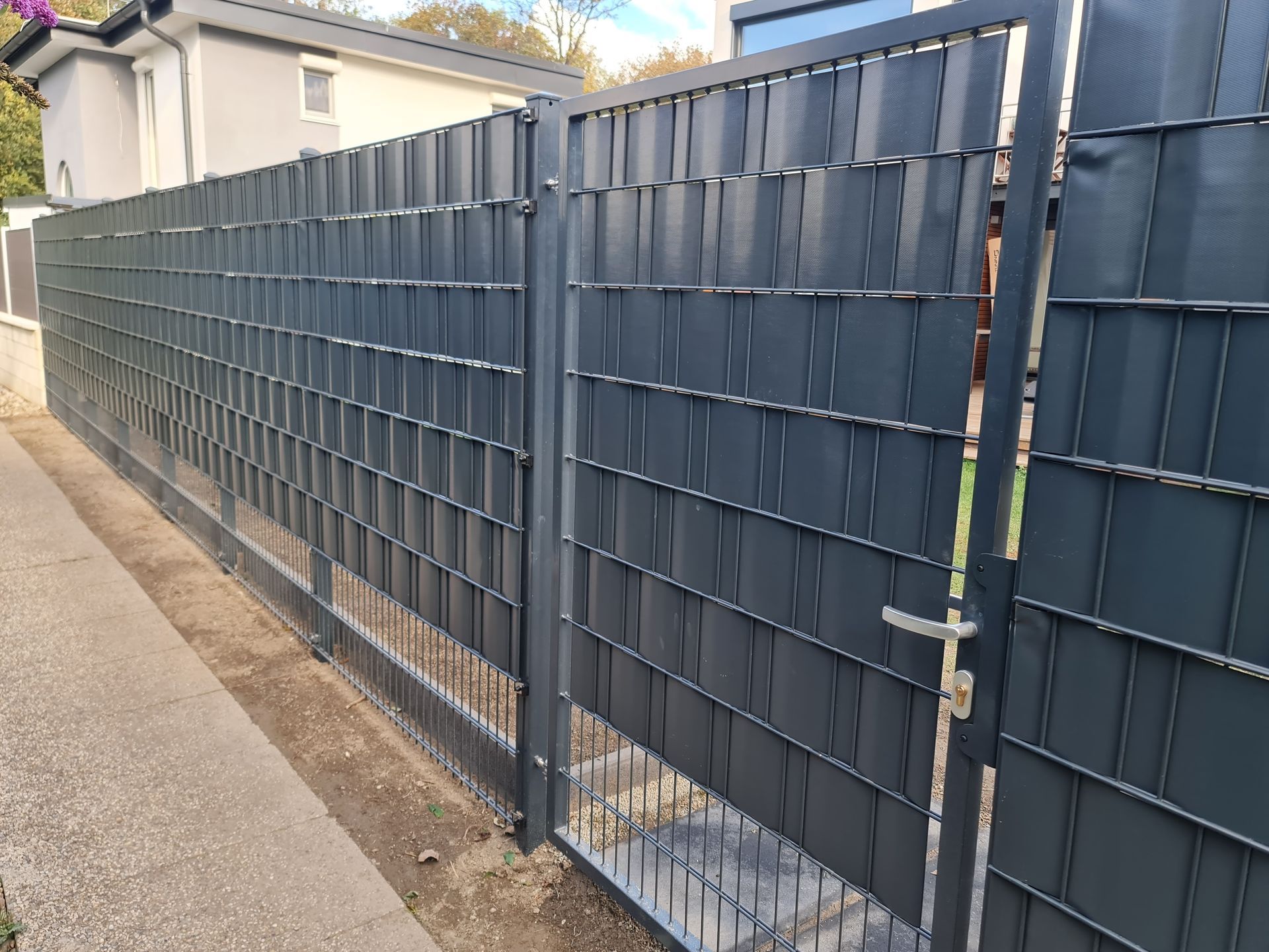 Graues Einfahrtstor aus Metall mit Zickzackmuster vor einem Haus mit grünen Hecken.