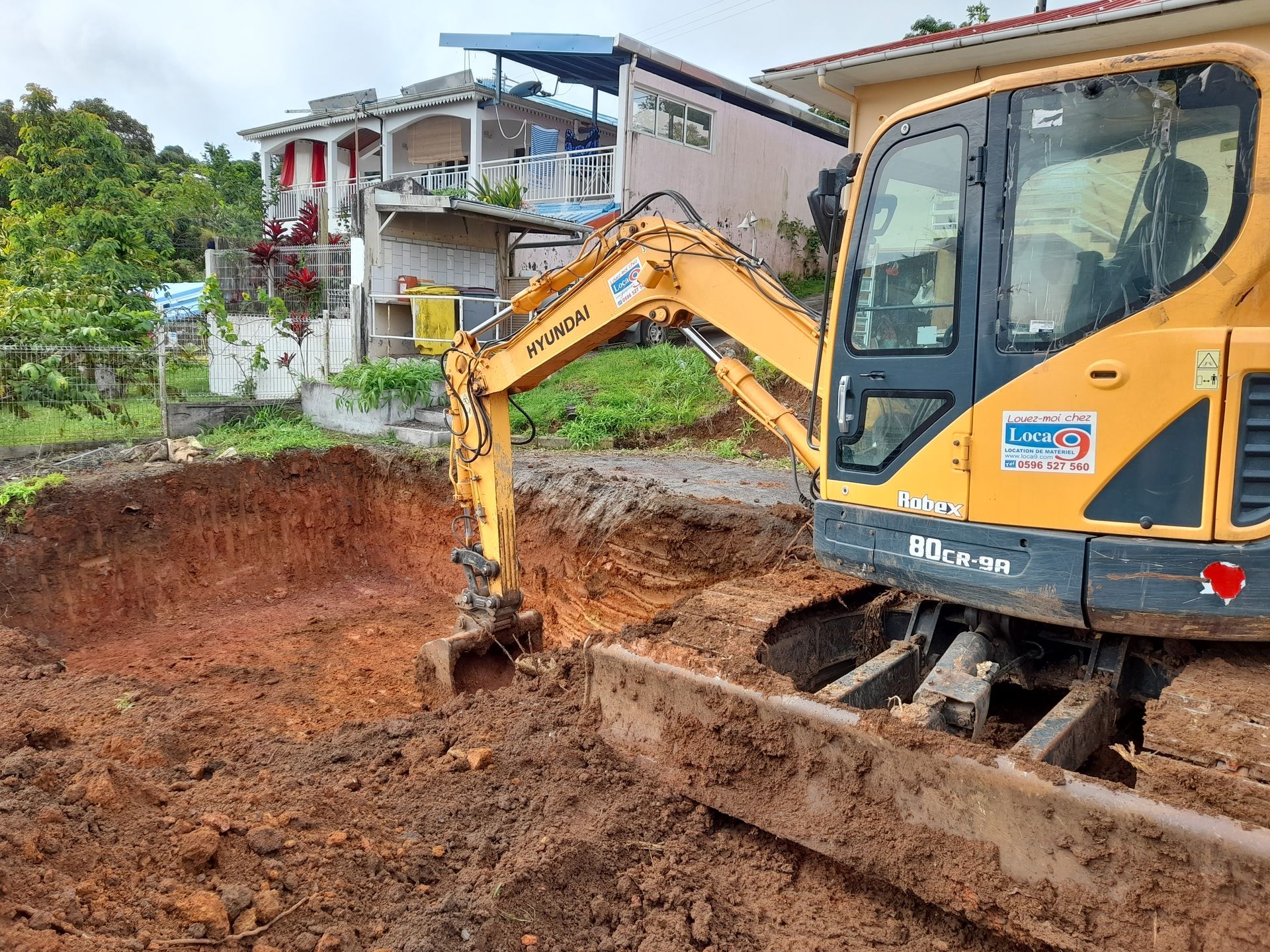 Une pelleteuse jaune creuse une tranchée boueuse à côté d'une maison dans une zone rurale.