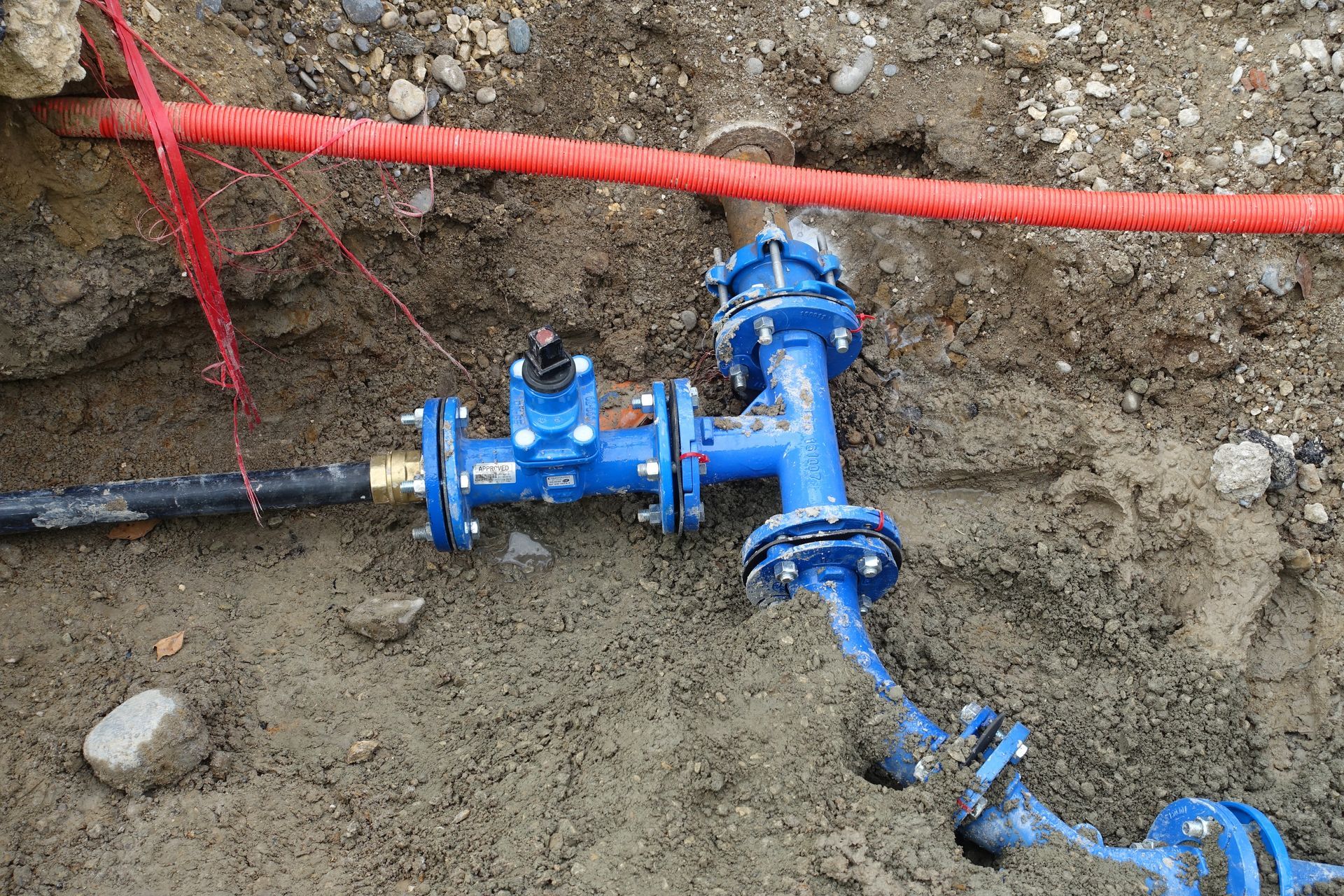 Ensemble de vannes de conduite d'eau bleue dans une tranchée de terre, avec des lignes rouges et noires au-dessus et une section de conduite qui fuit.