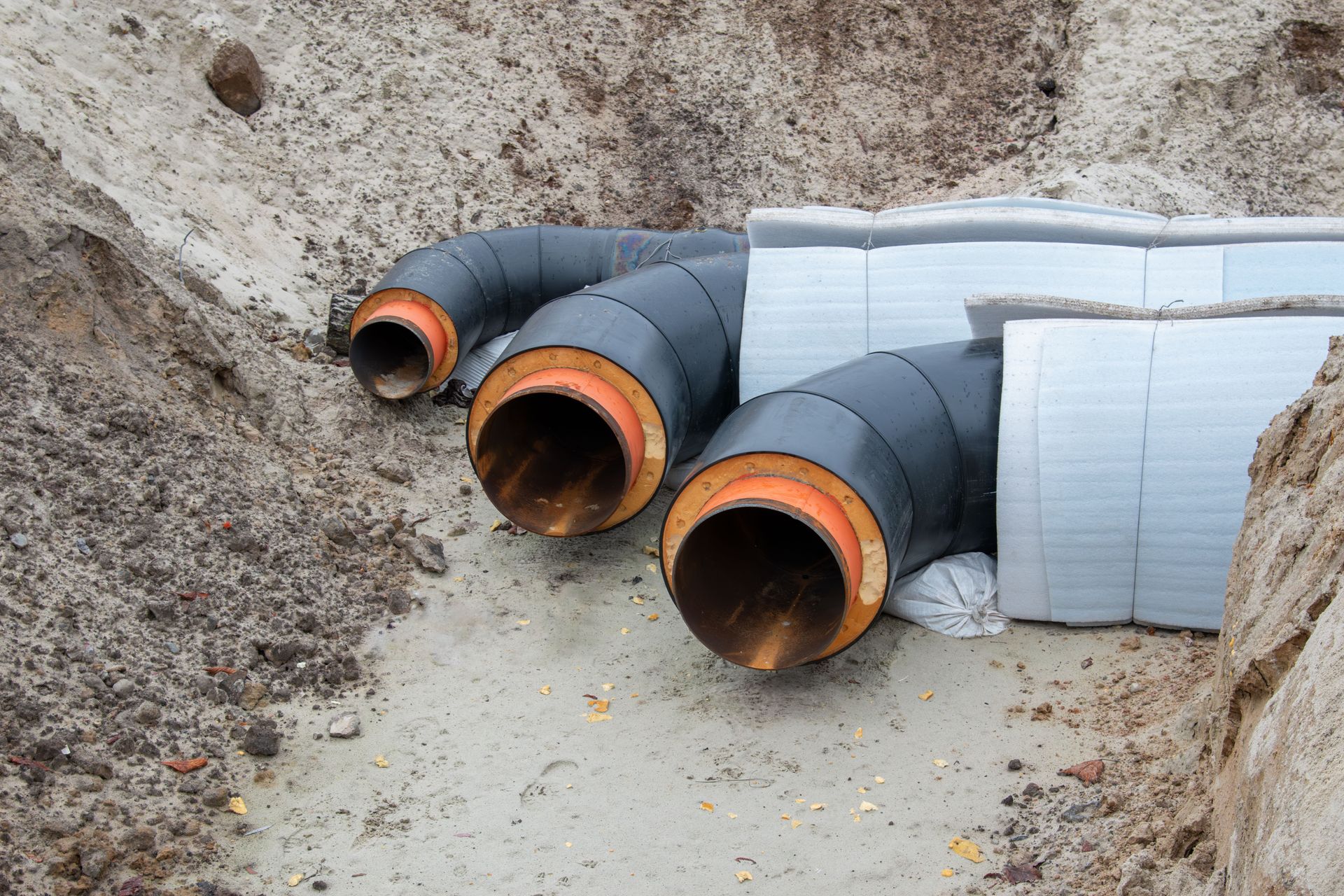 Des tuyaux de drainage noirs à bordures orange sont posés dans une tranchée à côté de blocs d'isolation emballés.