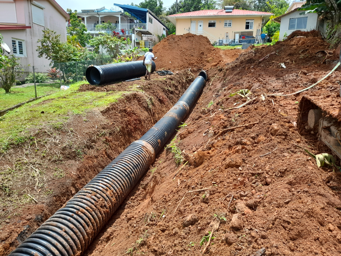 Tranchée de construction avec des tuyaux de drainage noirs posés dans la terre boueuse près des maisons.