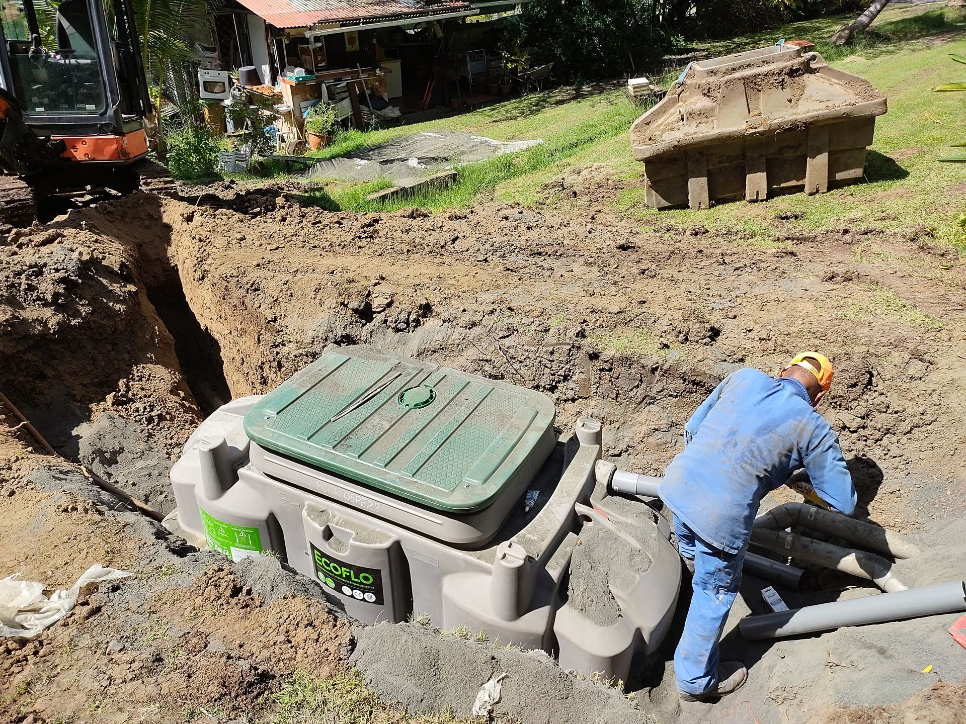 Un ouvrier se trouve près d'une cuve enterrée dans un chantier de fouilles boueux, avec des engins de chantier en arrière-plan.