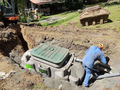 Un ouvrier se trouve près d'une cuve enterrée dans un chantier de fouilles boueux, avec des engins de chantier en arrière-plan.
