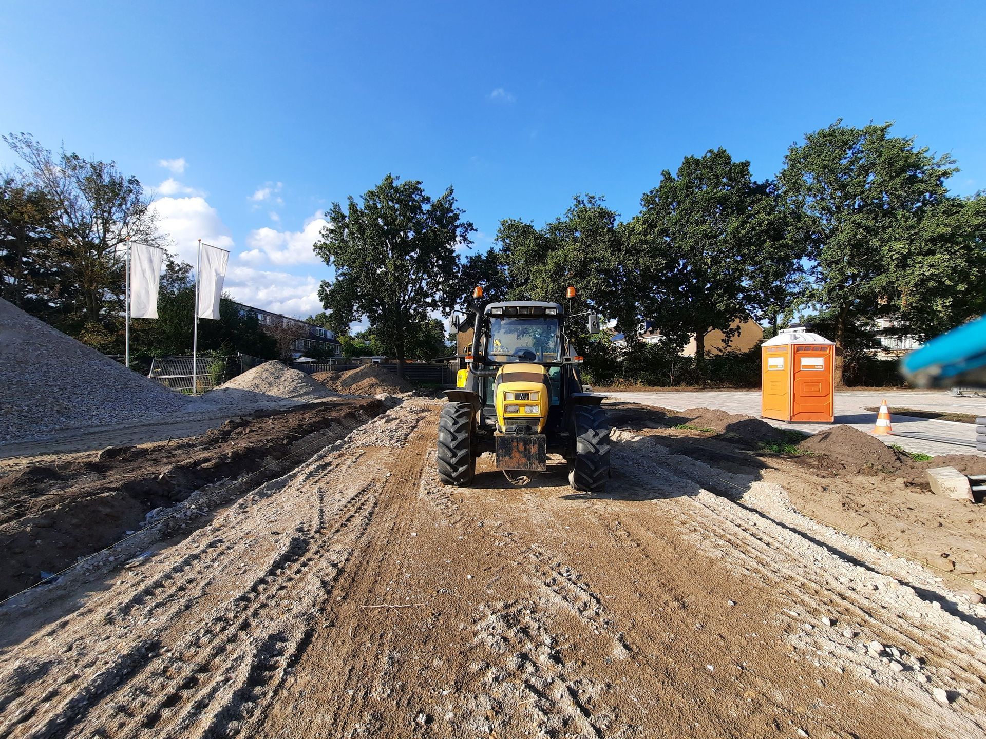 Chantier de construction avec un bulldozer jaune sur un chemin boueux, des tas de terre et des toilettes portables orange.