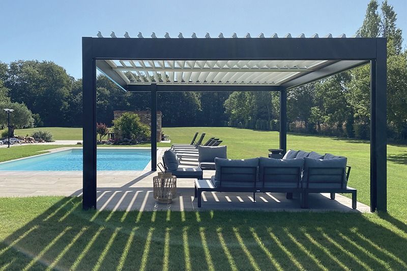 Une pergola à structure noire et à lames orientables au-dessus d'un salon de jardin gris.