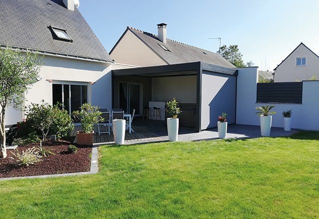 Patio arrière avec pergola moderne, table et plantes en pot, à côté d'une maison avec une pelouse verte.