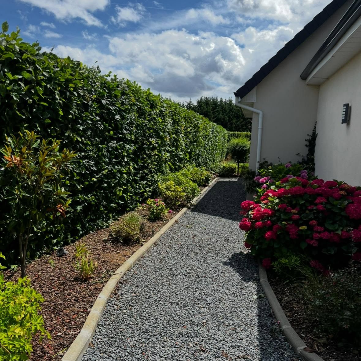 Chemin de gravier longeant une haie et une maison blanche, avec des fleurs roses et un ciel d'un bleu éclatant