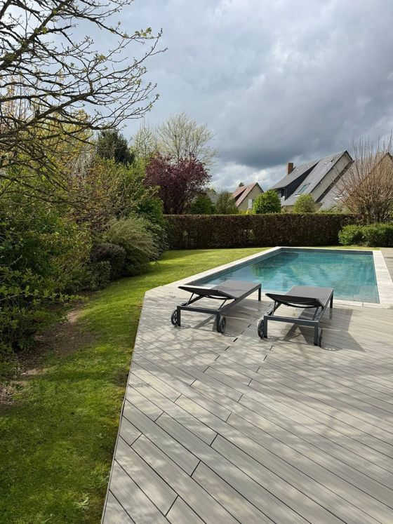 Terrasse avec chaises longues au bord d'une piscine turquoise, entourée de verdure sous un ciel nuageux.