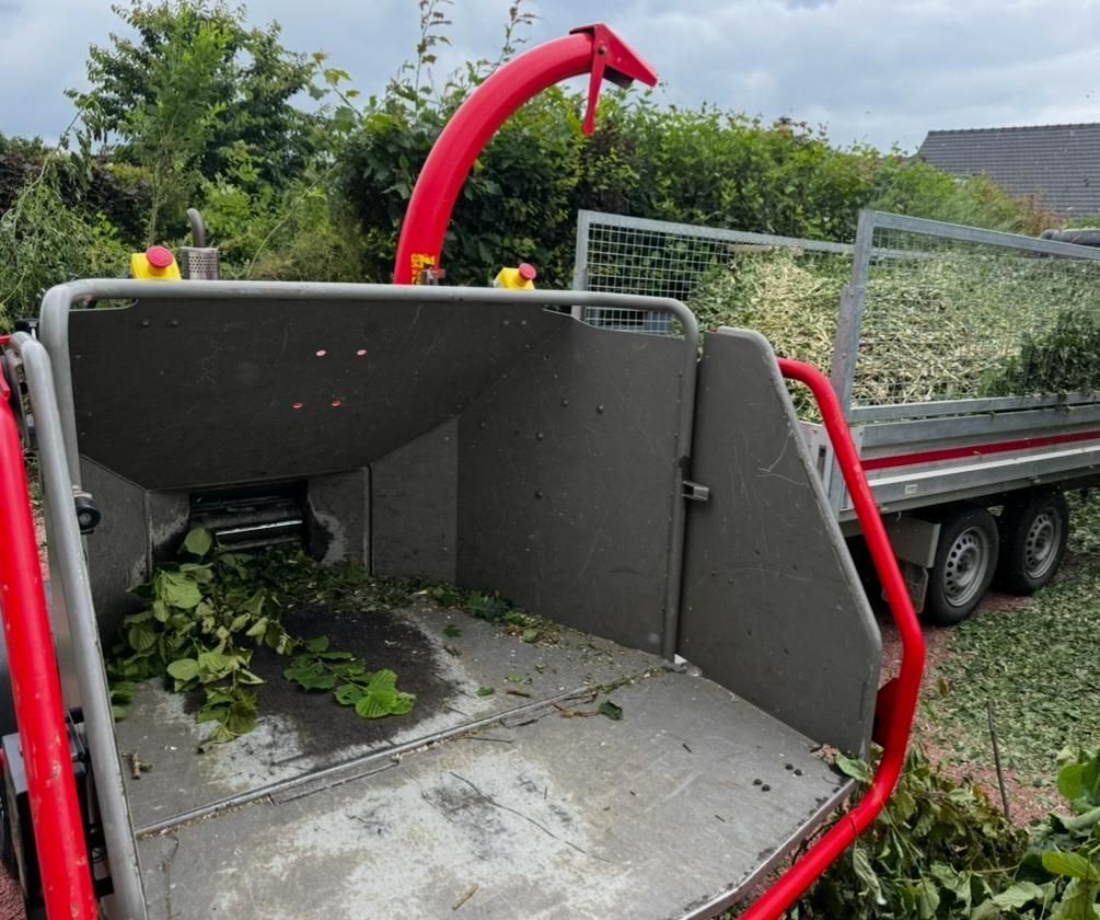 Broyeur de branches rouge à trémie ouverte, contenant des feuilles vertes, garé à côté d'un camion dans un jardin.