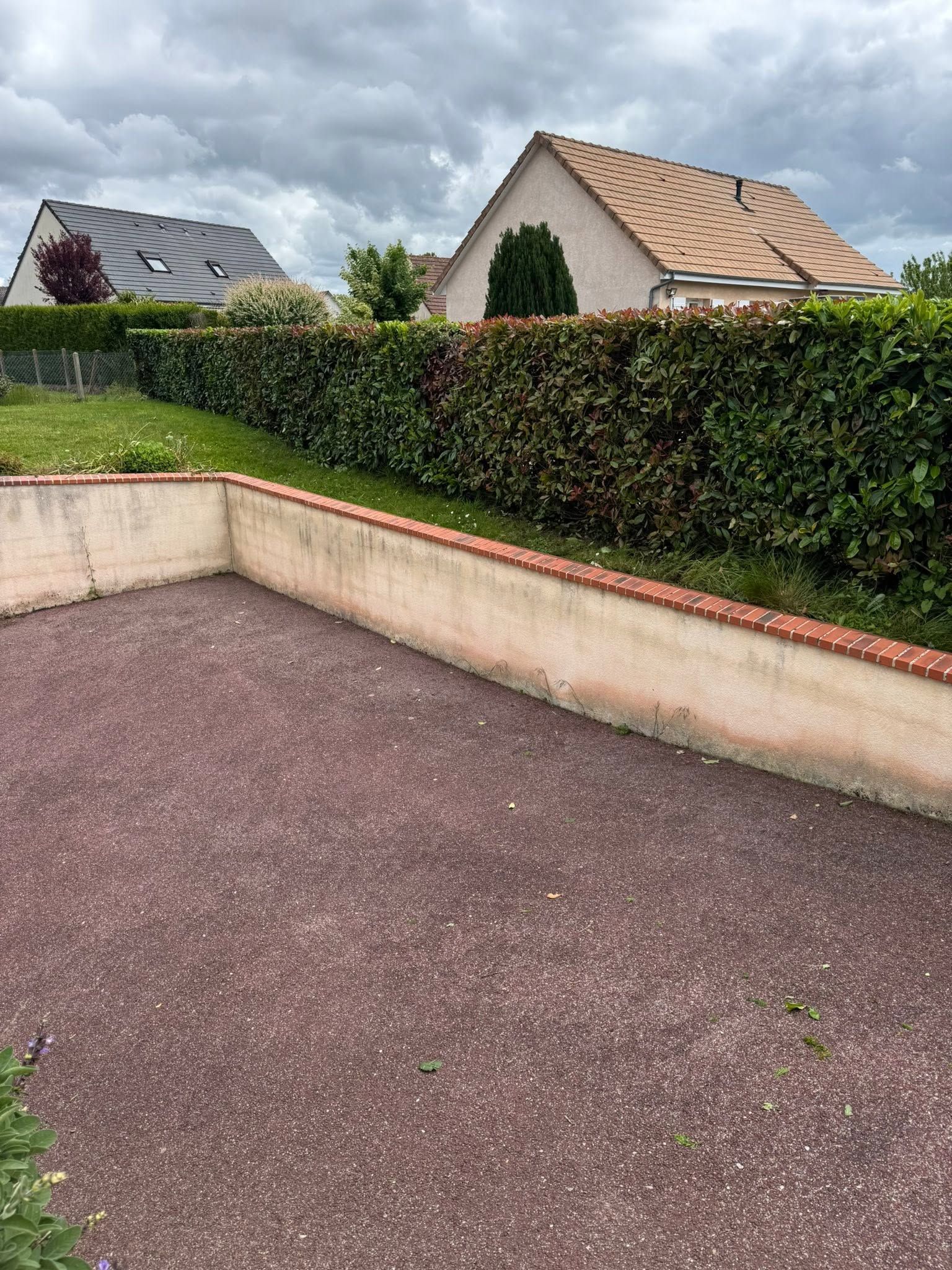 Terrasse arrière avec sol en gravier, haie et maisons voisines sous un ciel nuageux