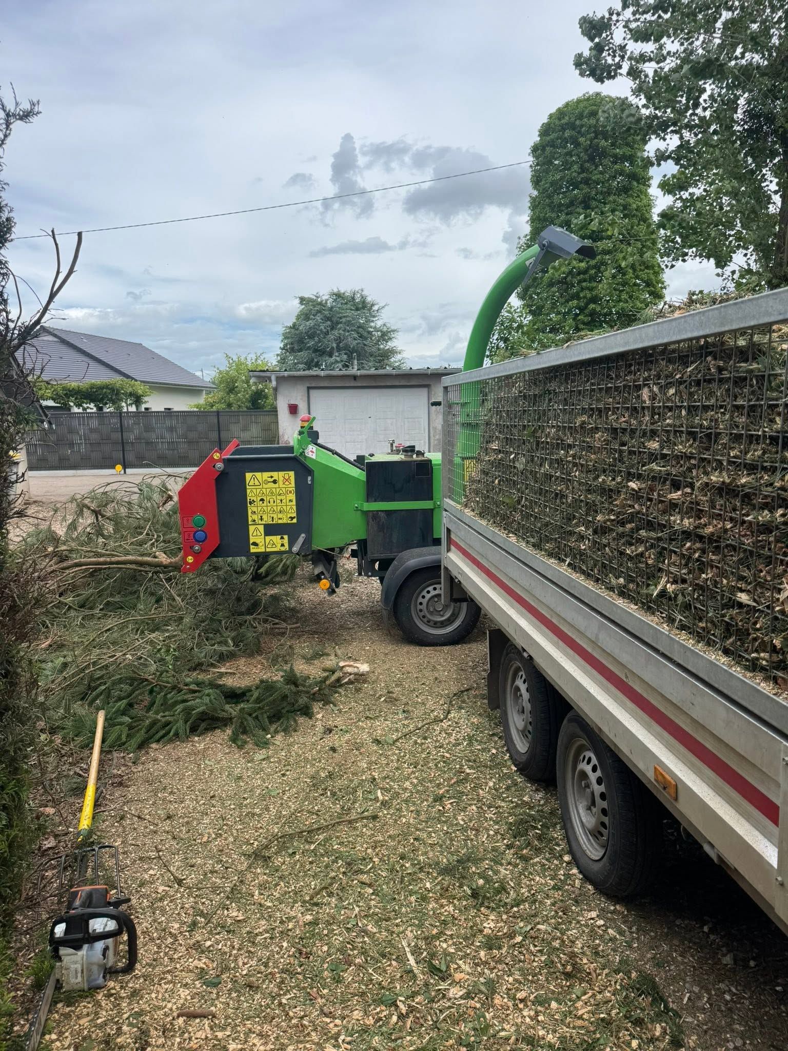 Une déchiqueteuse à bois verte, garée à côté d'un camion dans une cour, souffle des branches en un tas près d'une clôture.