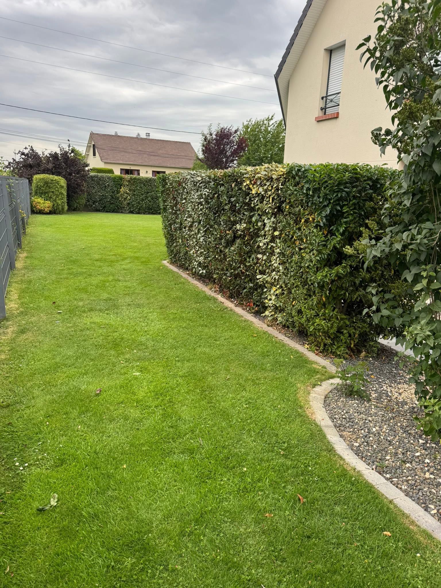 Pelouse verte bordée d'une haie et maison beige sous un ciel nuageux
