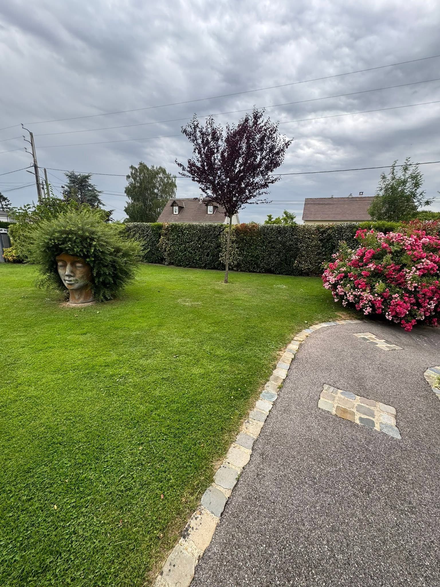 Jardin avec pelouse verte, haies taillées, buisson à fleurs roses et chemin de gravier sinueux sous un ciel nuageux