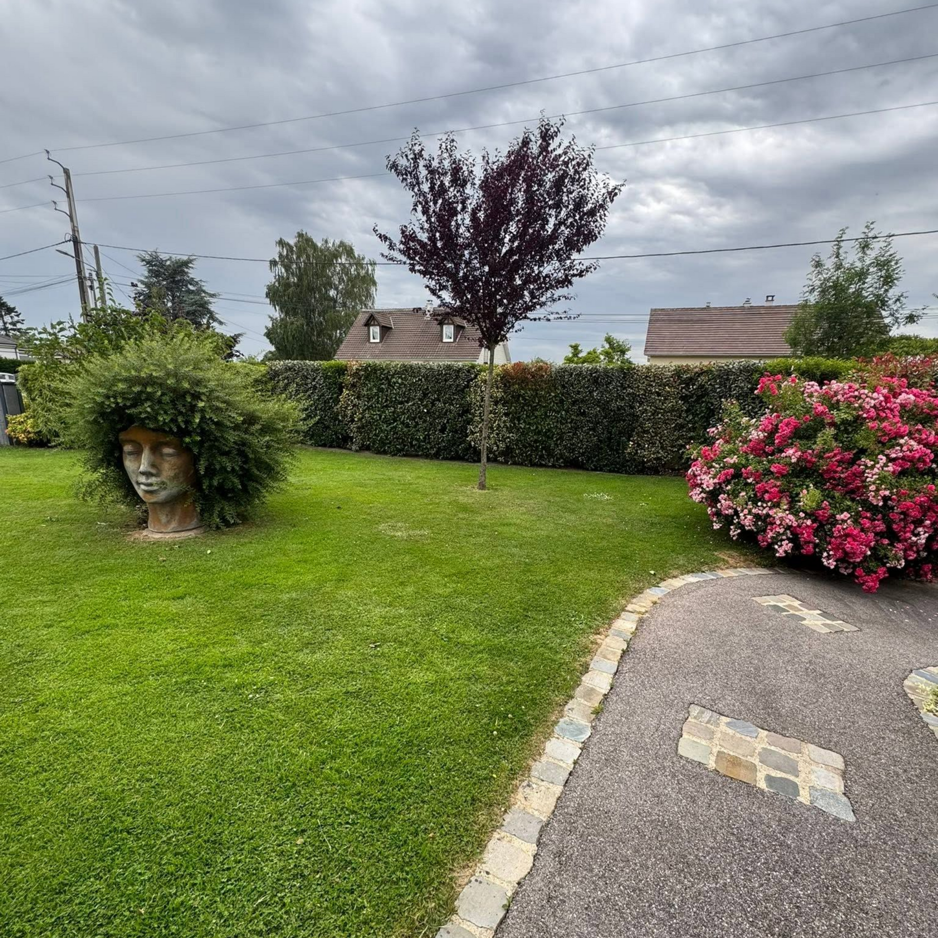 Pelouse de jardin avec jardinière sculptée en forme de visage, bordure de haie, fleurs roses et allée sinueuse sous un ciel nuageux.