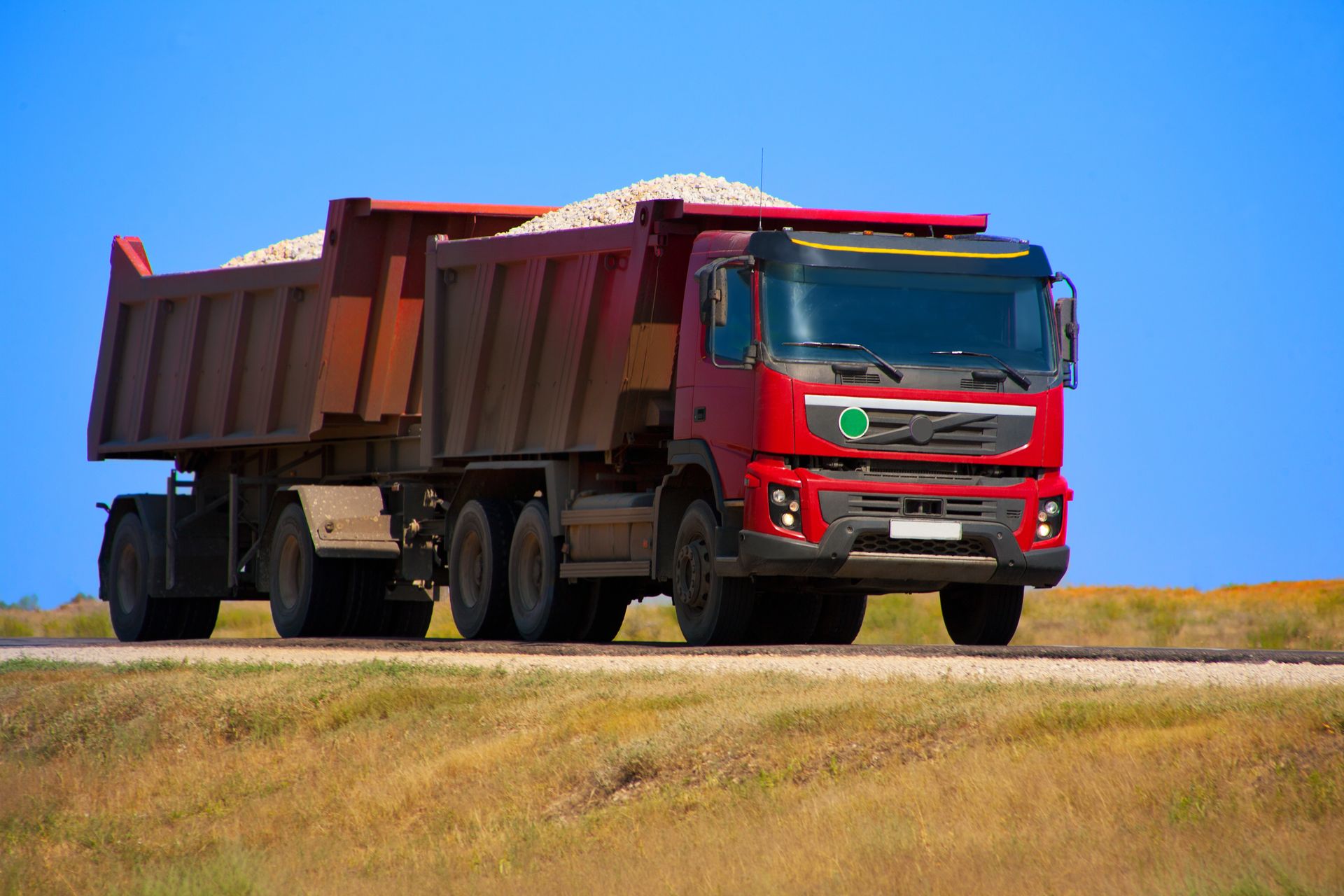 Camion à benne basculante rouge transportant du gravier sur une route, fond de ciel bleu.