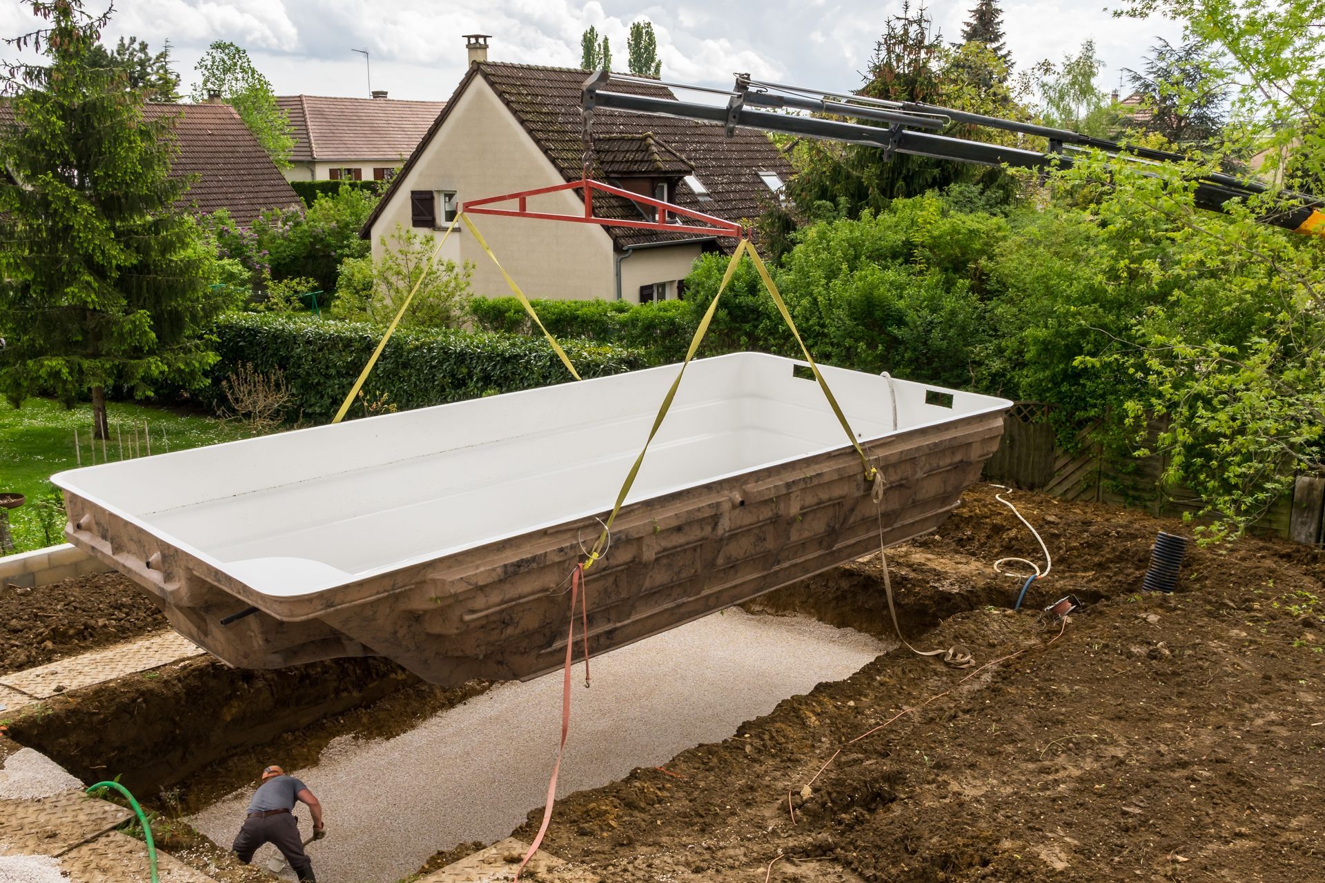 Une grande coque de piscine est soulevée par une grue dans une cour arrière boueuse, près de maisons et d'arbres.