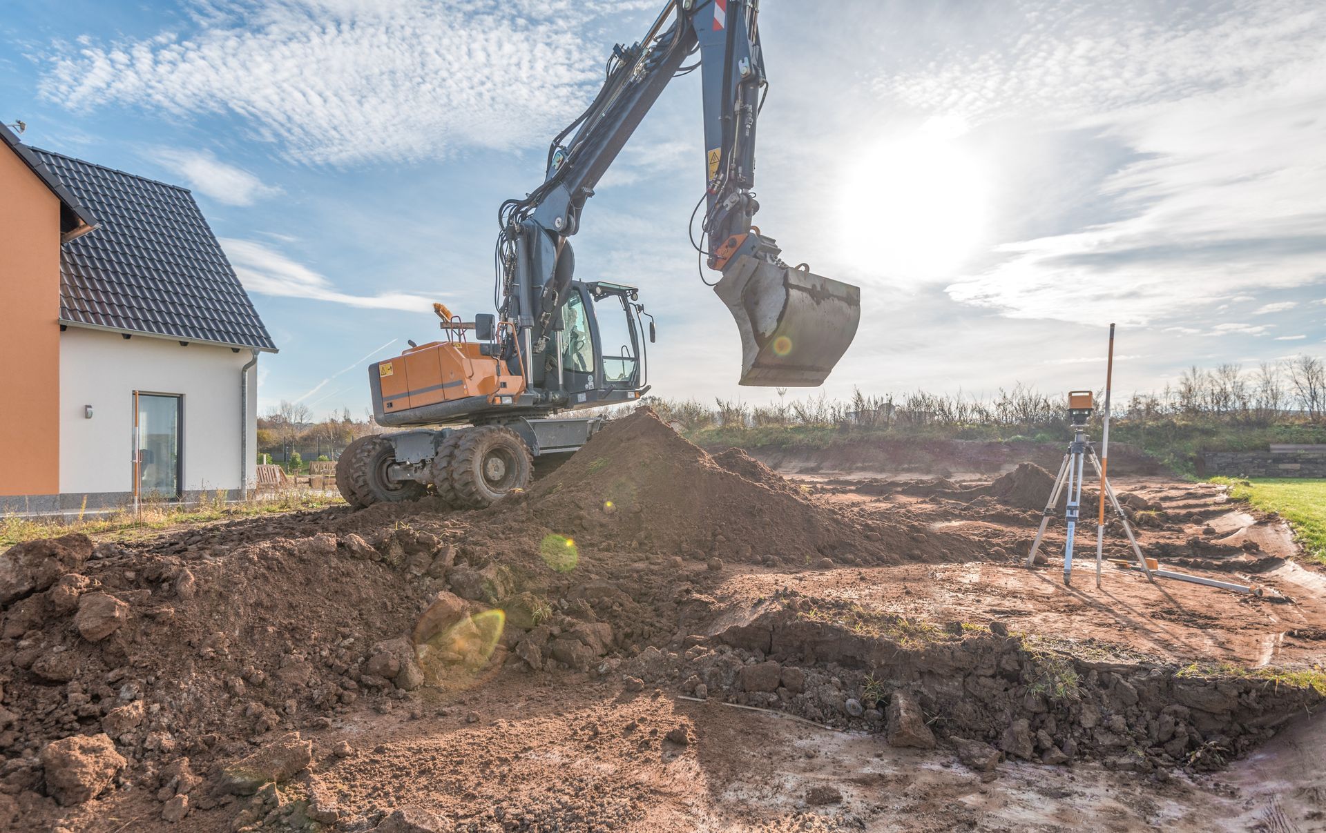Une pelleteuse creuse un monticule de terre à côté d'une maison sur un chantier de construction, sous un ciel radieux.