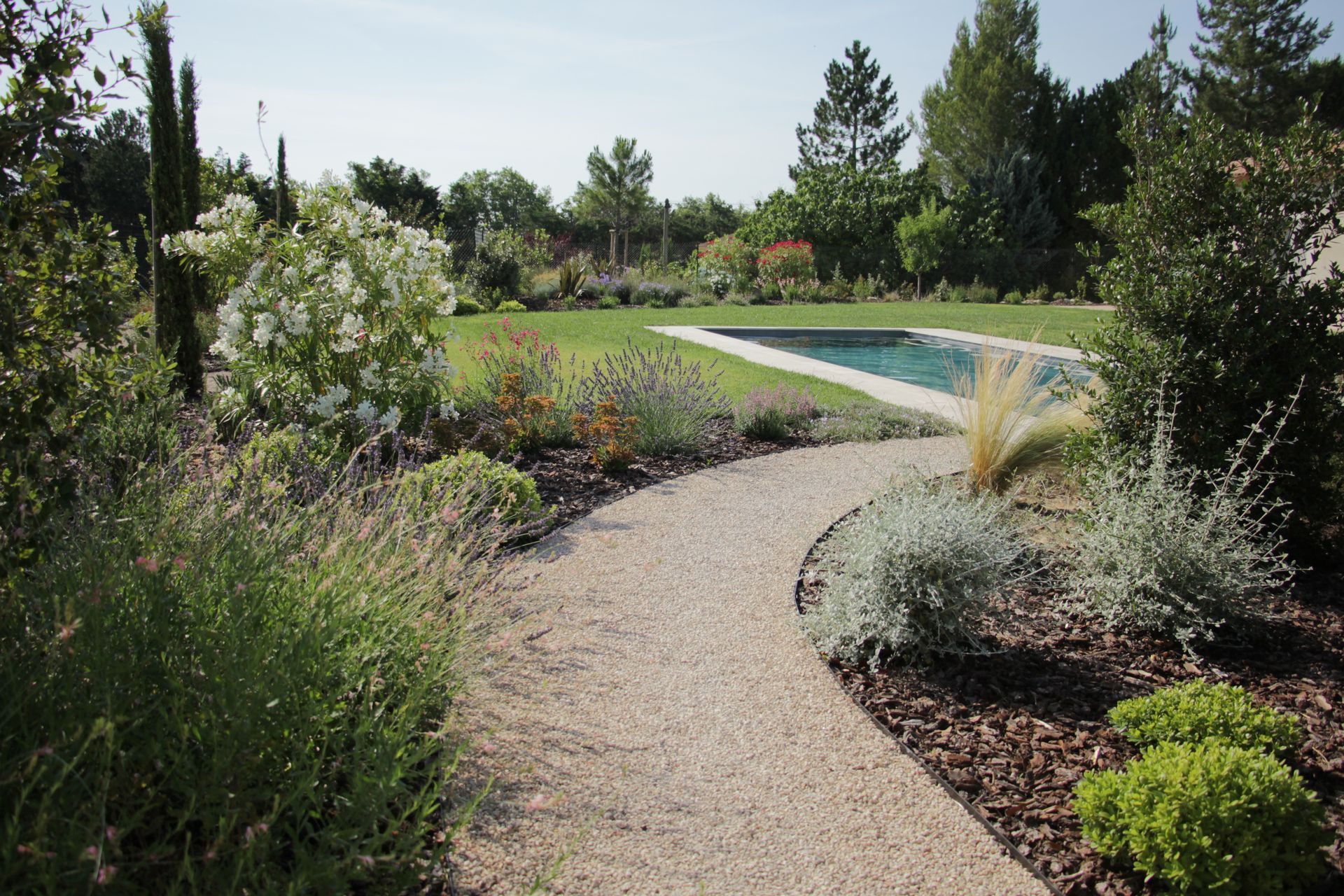 Chemin de gravier sinueux traversant un jardin paysager avec arbustes, arbres et piscine.