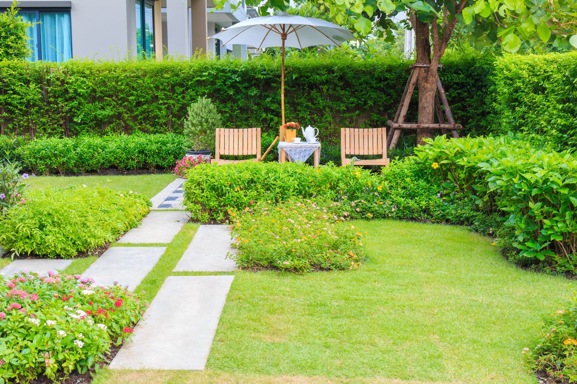 Jardin ensoleillé avec allée de pierres, haies taillées, pelouse et deux chaises sous un parasol.