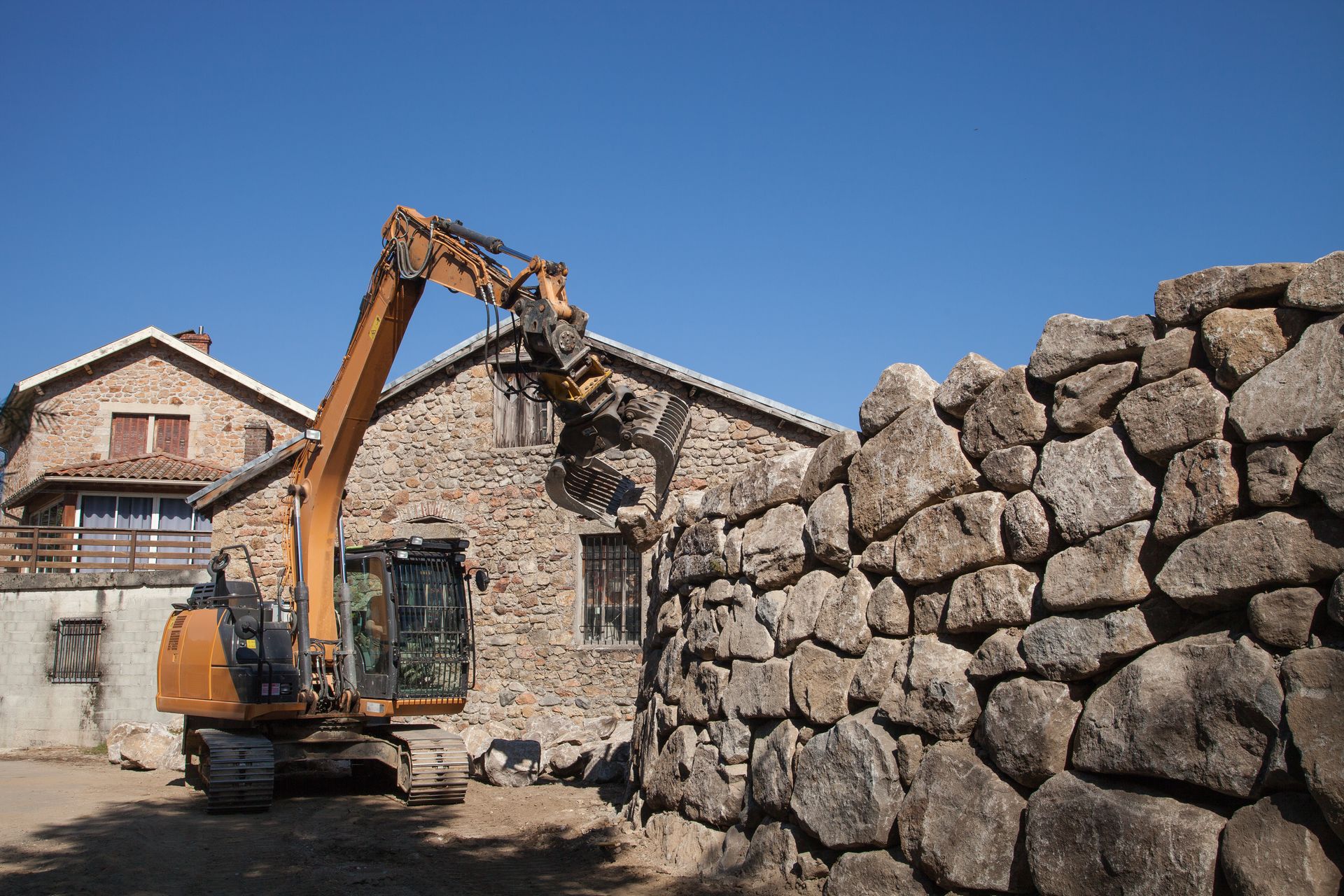 Une pelleteuse soulève une pierre pour la construction d'un mur de pierre à côté d'un bâtiment rustique sous un ciel bleu clair.