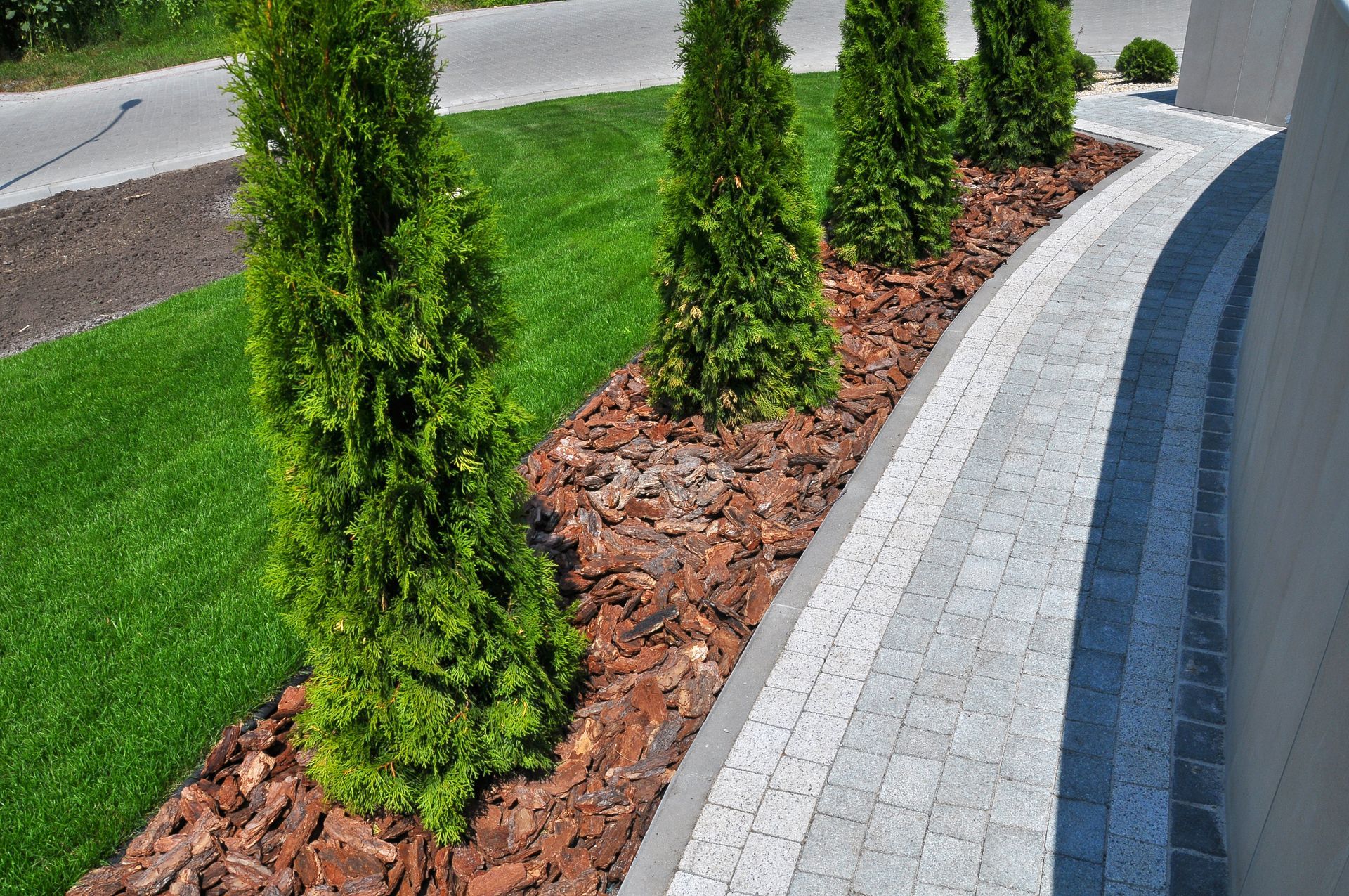 Allée paysagée sinueuse avec arbustes verts, paillis brun et bordure de pierres grises le long d'un chemin pavé.