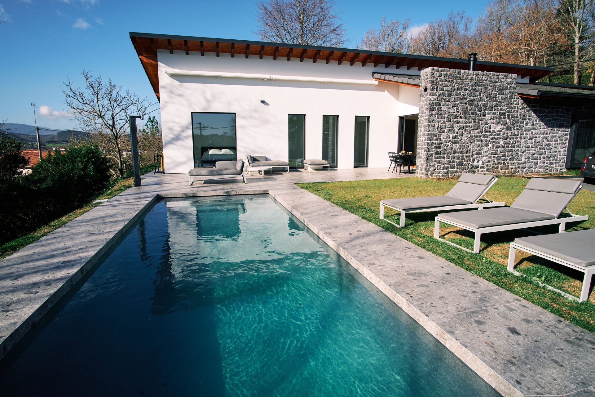 Maison moderne avec piscine, chaises longues et terrasse en pierre baignée de soleil.