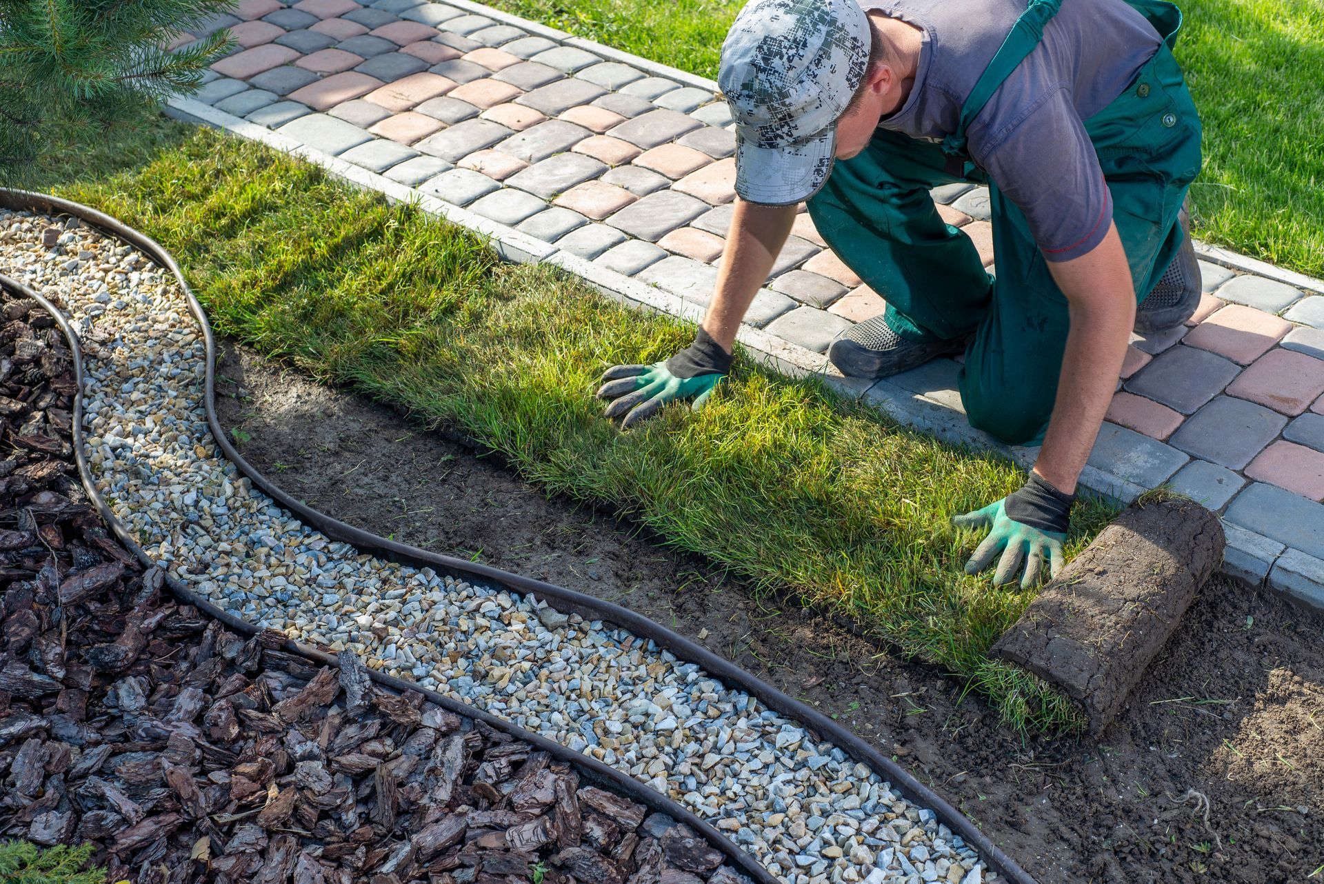 Une personne installe un rouleau de gazon le long d'une bordure de jardin, à côté d'une allée pavée.