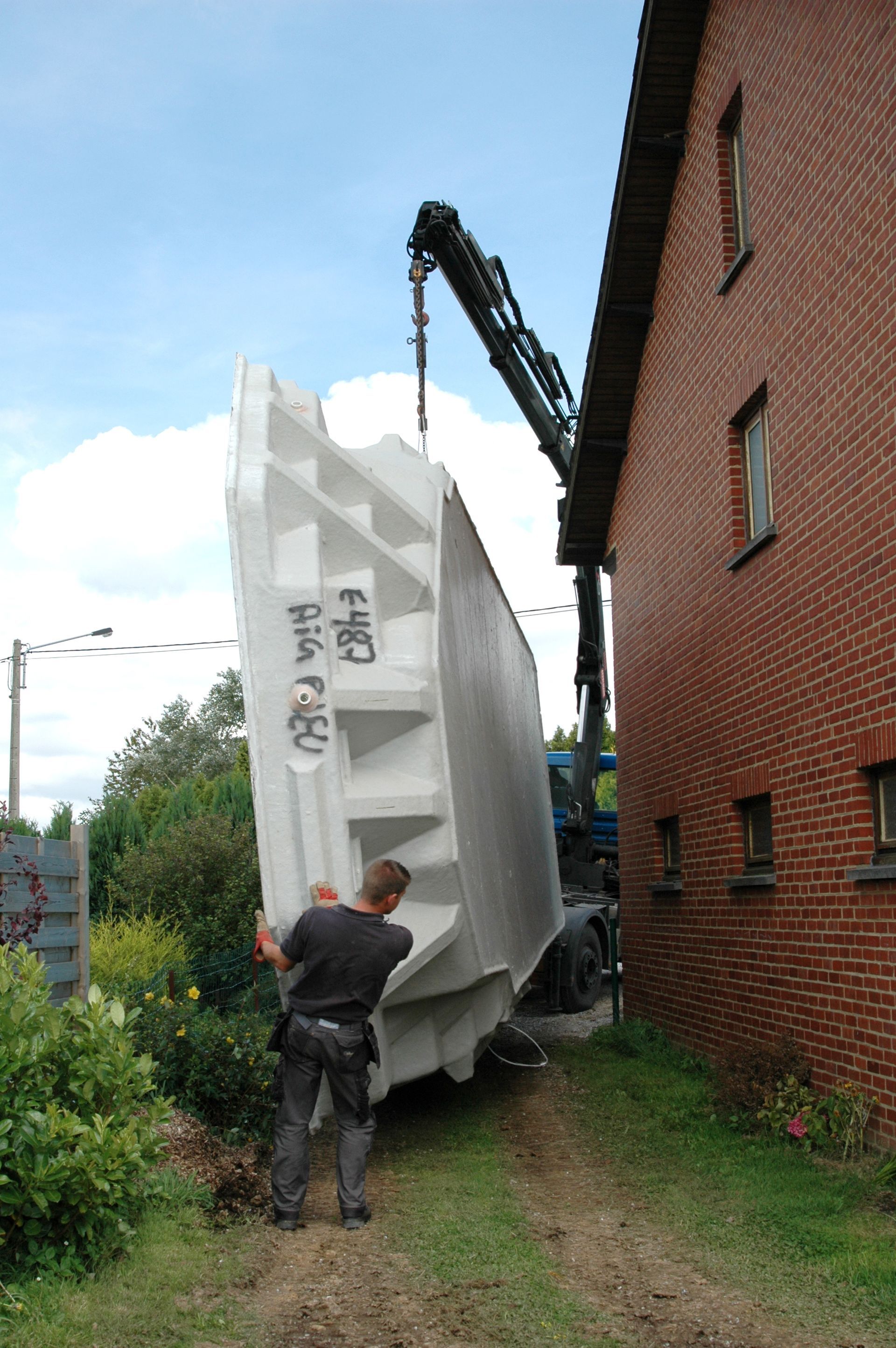 Un ouvrier guide une coque de piscine à côté d'un bâtiment en briques, soulevée par une grue dans une ruelle étroite.