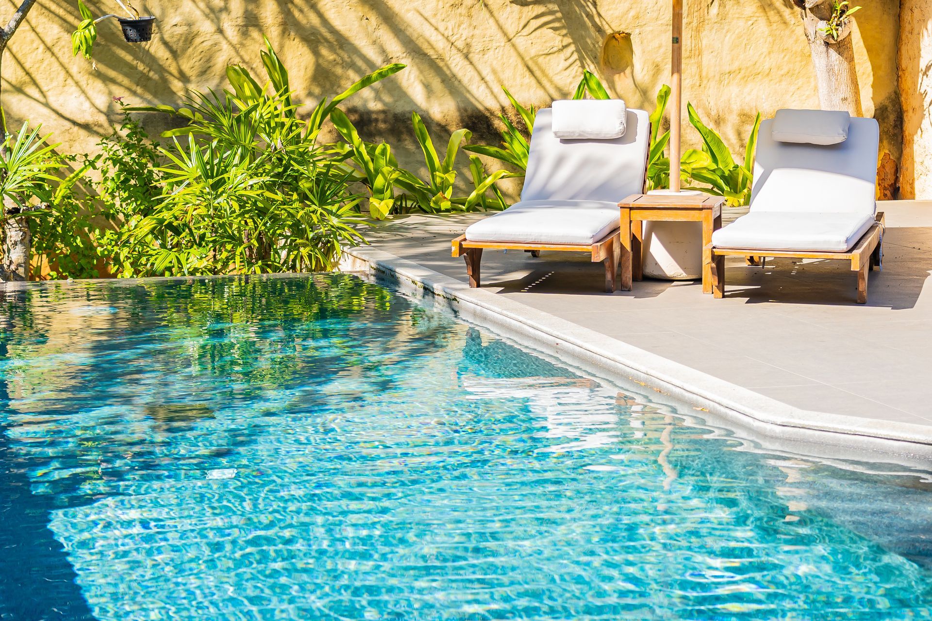 Piscine ensoleillée avec deux chaises longues blanches au bord d'une eau bleu clair et de végétation tropicale.
