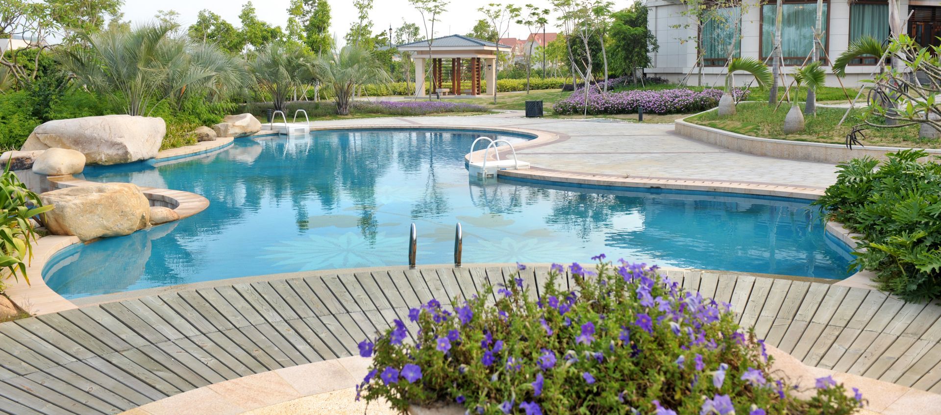 Piscine incurvée dans le jardin avec eau bleue, terrasse en pierre, chaises longues et fleurs violettes devant