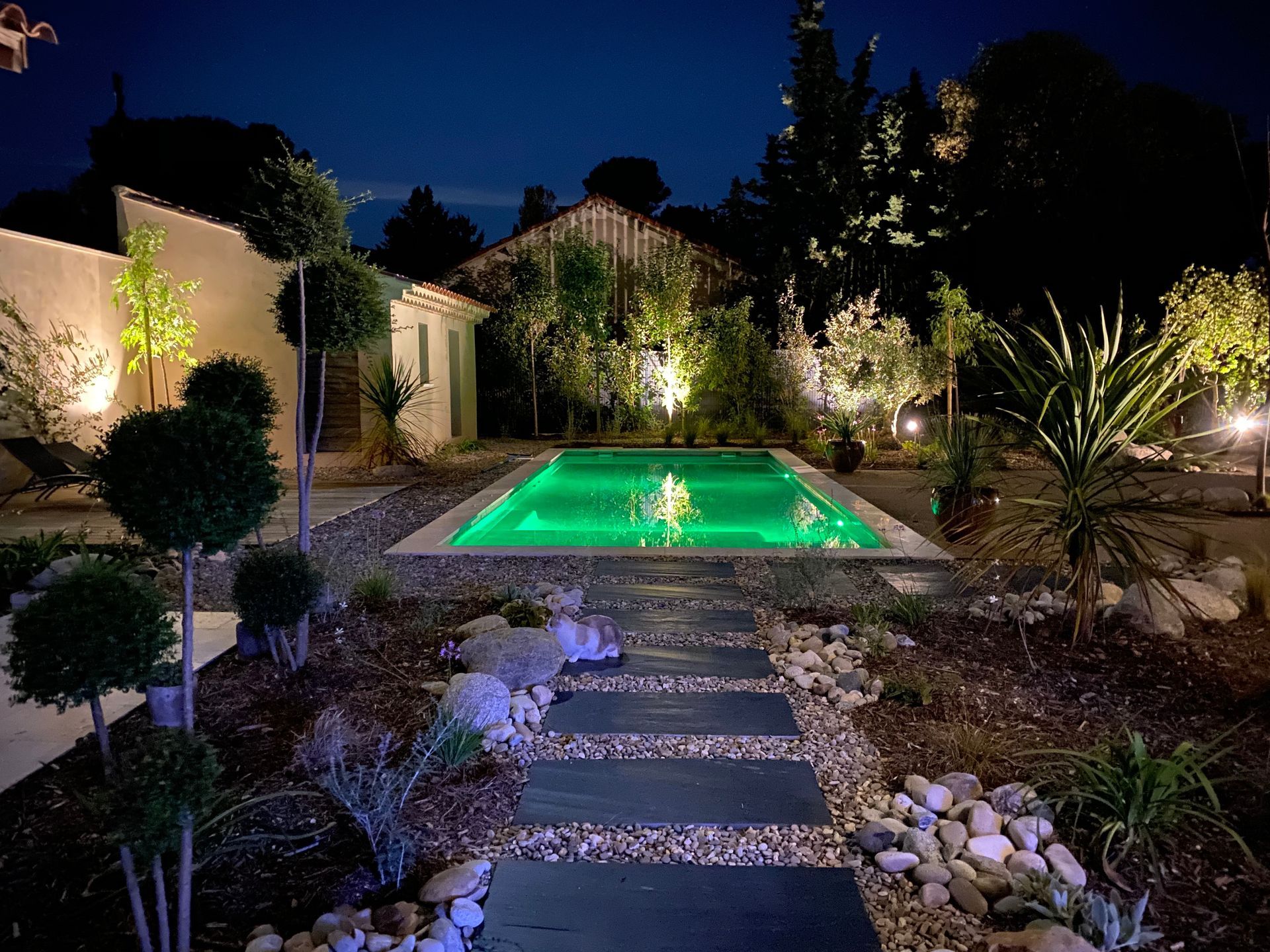 Patio arrière illuminé avec une piscine rectangulaire verte fluorescente, un chemin de pierre et un jardin paysager la nuit.