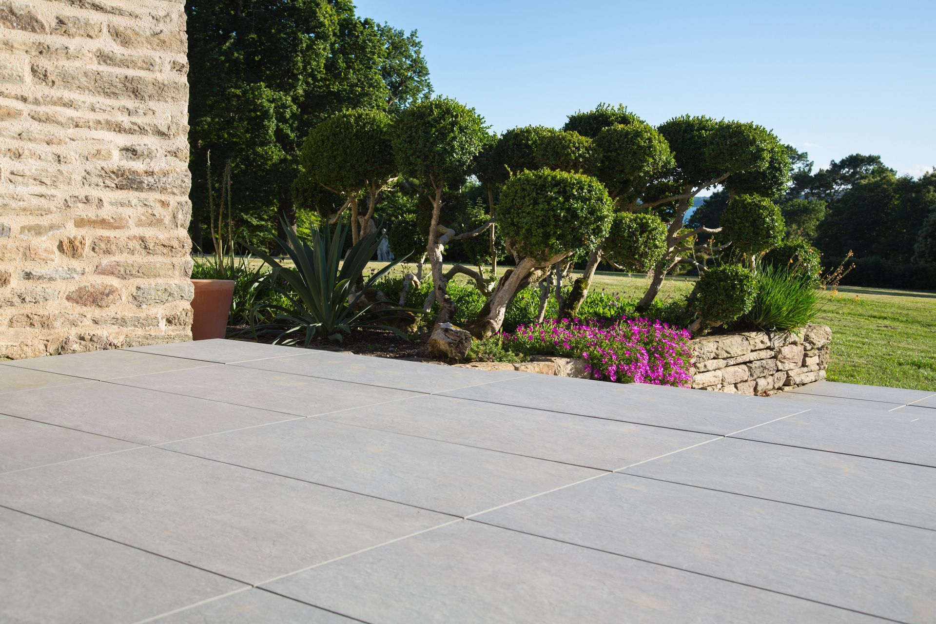Terrasse en pierre jouxtant un jardin paysager avec des arbustes taillés, des fleurs roses et un ciel d'un bleu limpide.