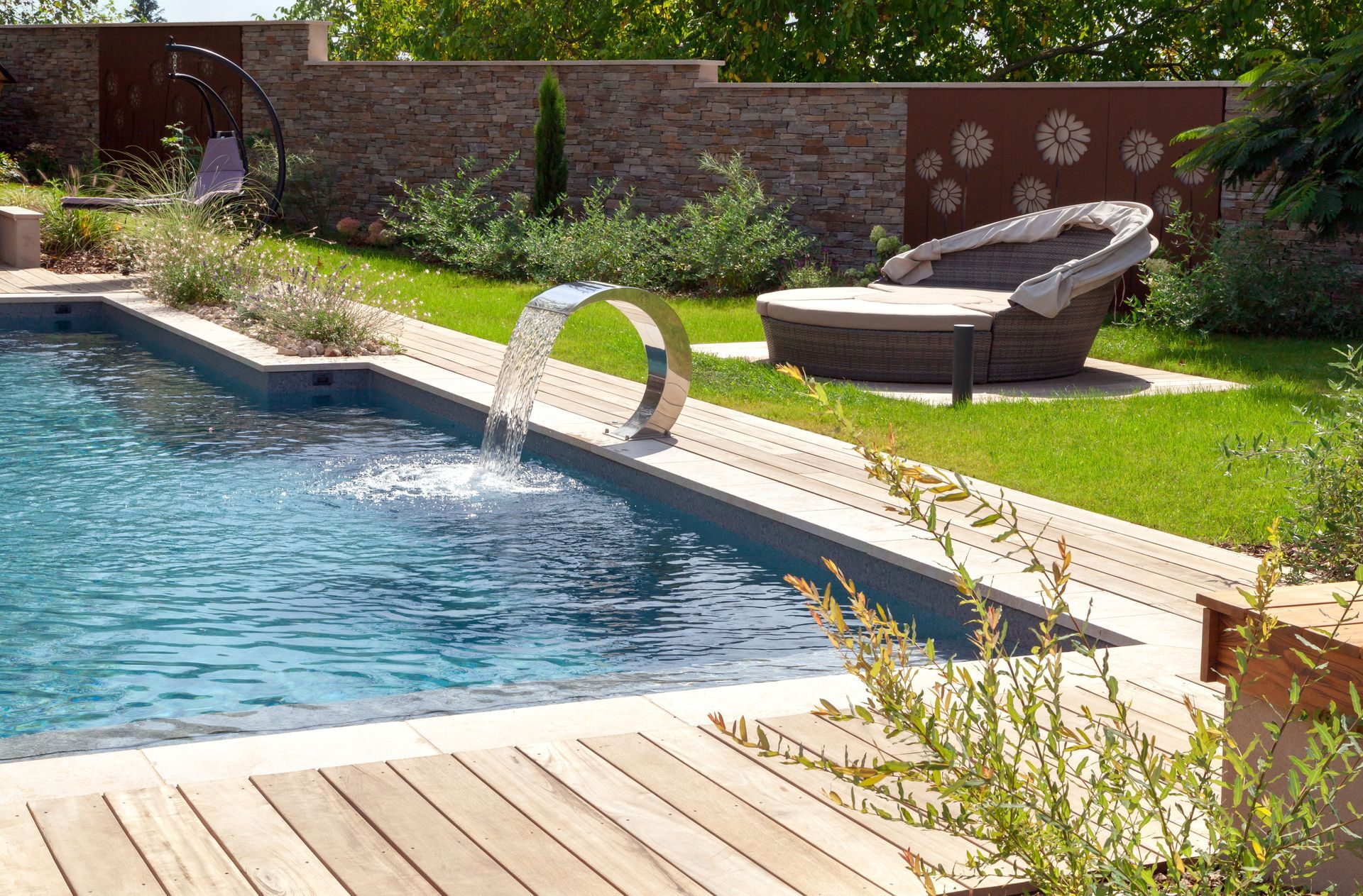 Piscine de jardin avec fontaine en arc de cercle, terrasse en bois, chaise longue et pelouse verdoyante.