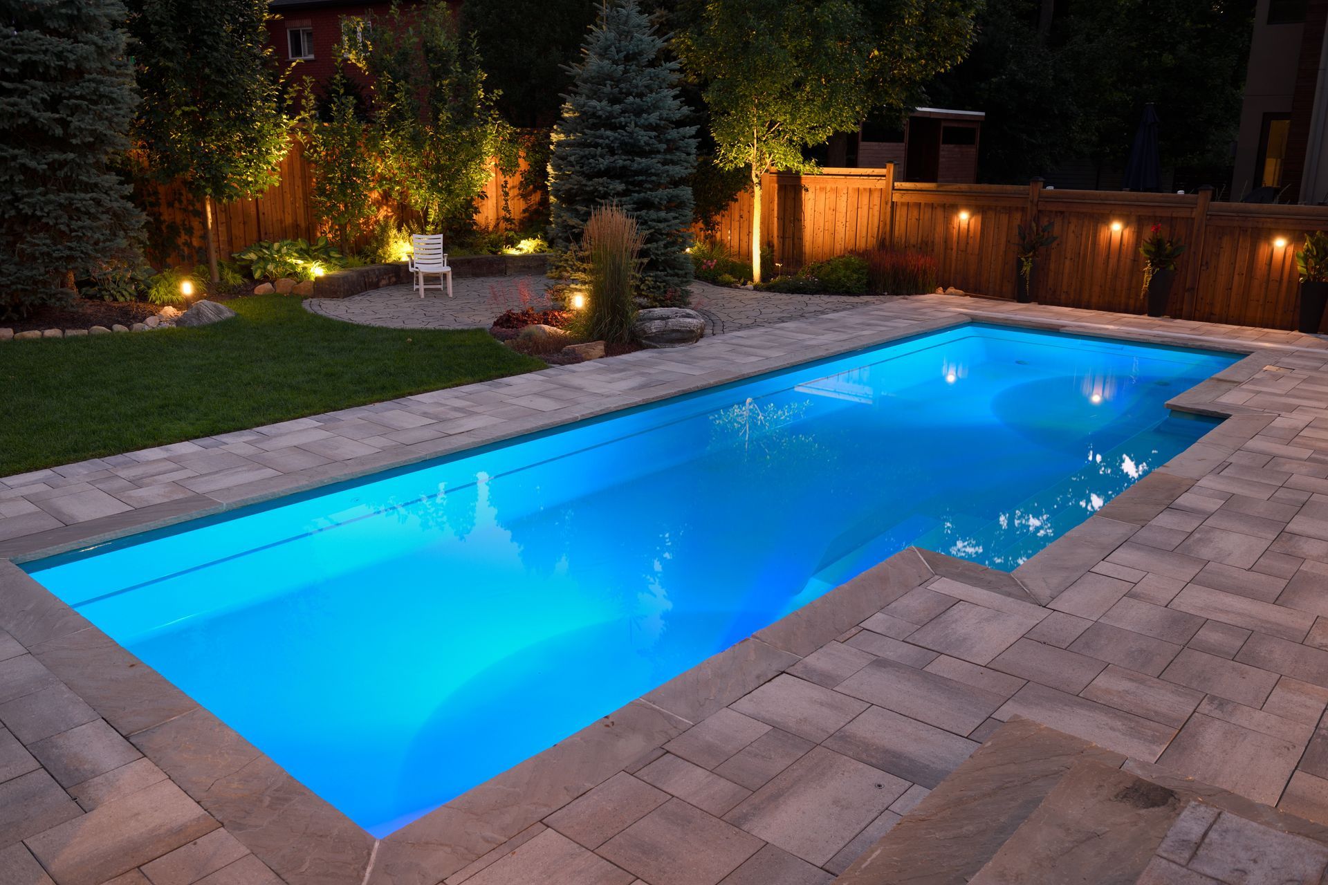 Piscine illuminée dans le jardin, aux reflets bleus la nuit, bordée d'une terrasse en pierre et de luminaires paysagers.