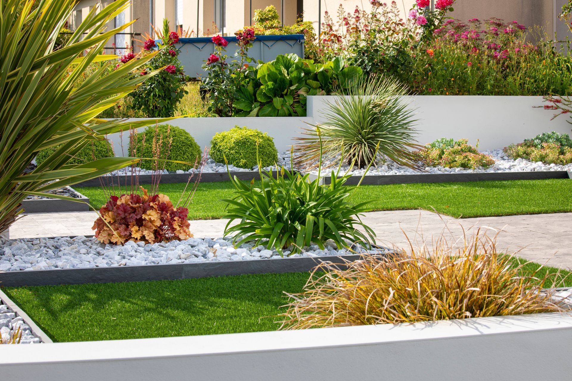 Cour moderne avec parterres de fleurs sur plusieurs niveaux, pelouse verte et plantes ornementales dans des jardinières.