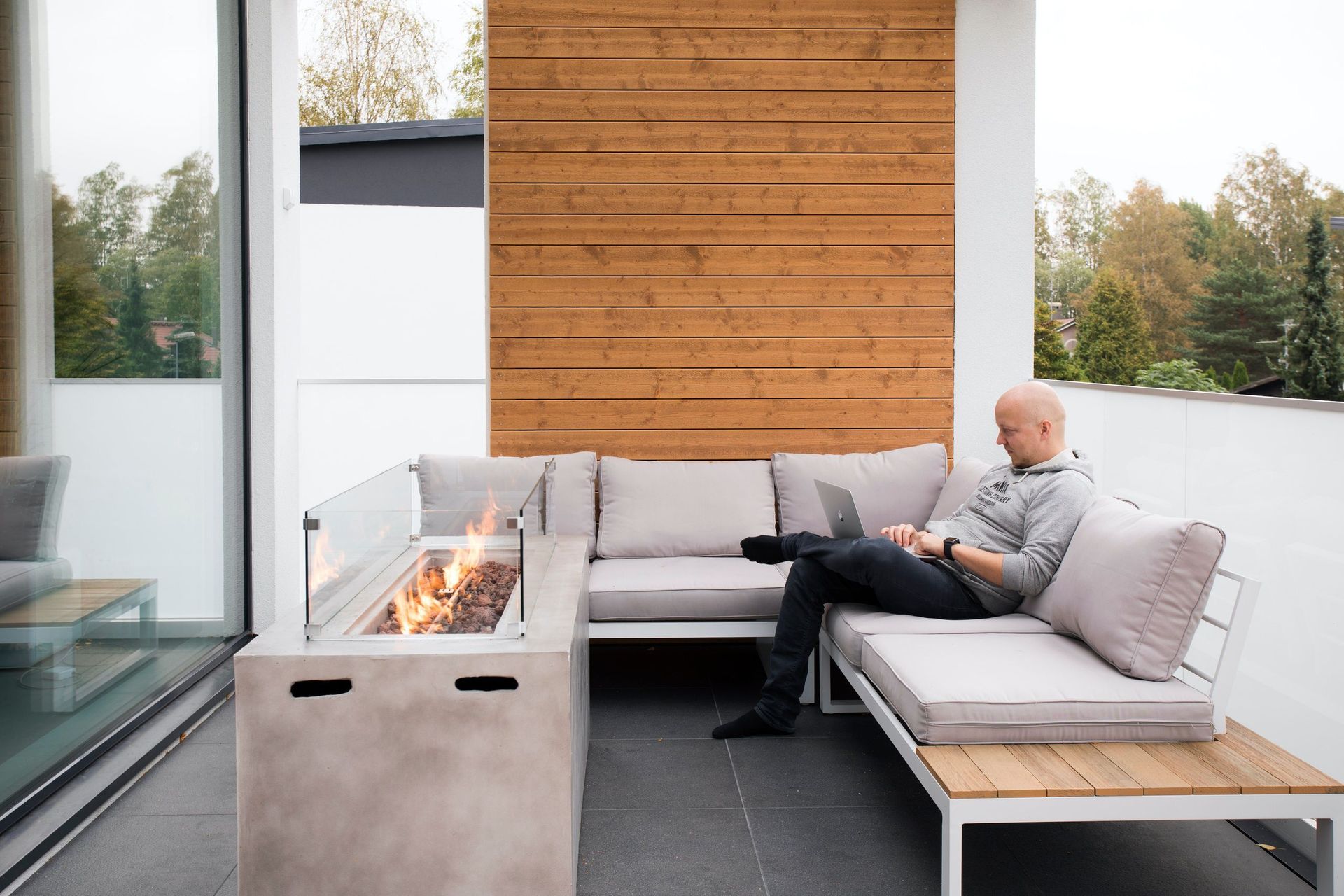 Moderni patio, jossa on nuotiopaikka ja harmaita sohvia puumuurin vieressä; henkilö istuu rentoutuen puhelimen kanssa.